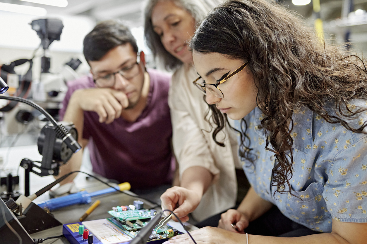 College instructor assisting electrical engineering students. Close view of mature female educator standing between early 20s classmates and offering guidance as they work on project.